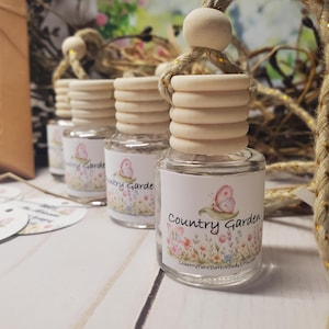 May include: A clear glass car air freshener bottle with a light wood cap and a hanging jute rope. The label features a pink butterfly, colorful wildflowers, and the text Country Garden and CountryPureBathNBodyEtsy.com in black script.