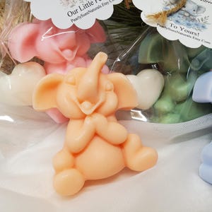 May include: Several elephant-shaped soaps in various colors, including peach, pink, green, and blue. Each soap has a heart-shaped accent. The soaps are packaged in clear cellophane bags with decorative tags. The tags read "Our Little Flower" and "To Yours!"