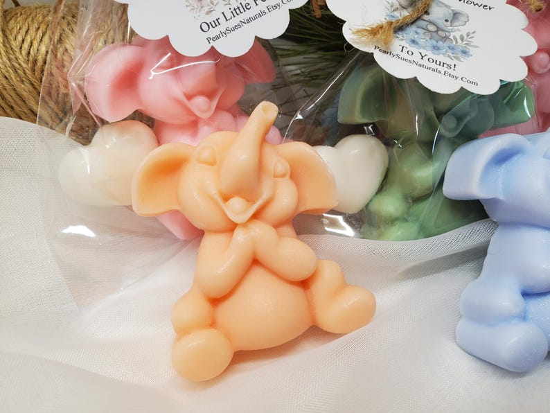May include: Several elephant-shaped soaps in various colors, including peach, pink, green, and blue. Each soap has a heart-shaped accent. The soaps are packaged in clear cellophane bags with decorative tags. The tags read "Our Little Flower" and "To Yours!"