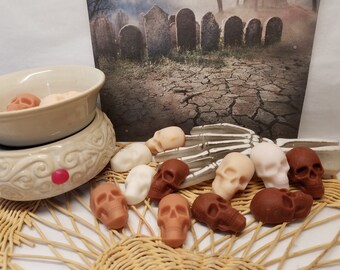 May include: A collection of skull-shaped wax melts in various shades of brown, white, and pink. The melts are arranged on a woven wicker surface with a white ceramic wax warmer and a skeletal hand in the background.