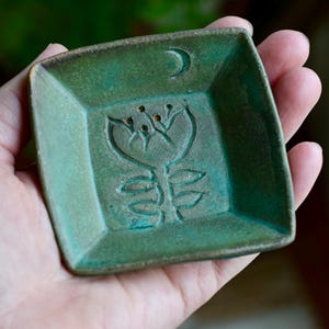 Può includere: Un piatto quadrato in ceramica con smalto verde. Il piatto presenta un motivo floreale e una luna crescente. Il piatto è tenuto in mano, mostrando le sue dimensioni e la sua consistenza. Il piatto viene probabilmente utilizzato per contenere piccoli oggetti.