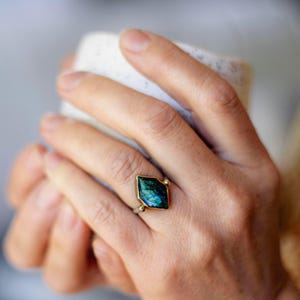 May include: A ring featuring a kite-shaped, iridescent blue-green labradorite gemstone set in a gold-toned metal bezel. The ring band is thin and metallic, with small gold-toned spheres accenting the base of the stone setting.