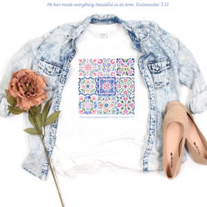 Puede incluir: Camiseta blanca con un vibrante diseño floral de azulejos en tonos rosa, azul y verde. La camiseta incluye el texto "He has made everything beautiful in its time. Ecclesiastes 3:11". Una chaqueta vaquera azul claro y bailarinas beige también están presentes.