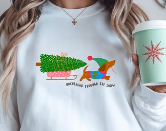 Peut inclure: Sweat-shirt blanc avec un motif festif. Le motif représente un teckel tirant un sapin de Noël sur une luge rose, avec le texte "Dachshund Through The Snow". Le chien porte un bonnet et un pull bleu et vert.