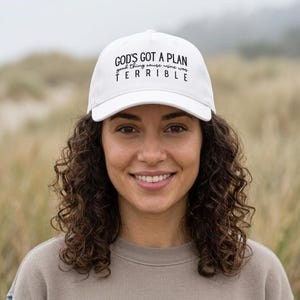 May include: A white baseball cap featuring black text printed on the front panel. The text reads GOD'S GOT A PLAN, followed by the smaller script text good thing cause wine was, and the word TERRIBLE in large capital letters at the bottom.