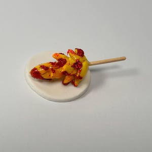 Può includere: Un lecca-lecca di caramelle giallo e arancione con glitter rossi, su una base circolare bianca e un bastoncino di legno. La caramella ha la forma di un fiore o di una stella. Lo sfondo è bianco.