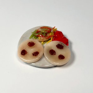 May include: Miniature ceramic plate with two round, light-colored food items, each with three dark red spots. The plate also holds a colorful salad with red, green, and yellow vegetables, and a dollop of red sauce. The food items are arranged on a white surface.