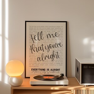 May include: A framed print with the words "tell me that you're alright" in a cursive font. A record player with a vinyl record is on a wooden shelf, next to a white and orange globe lamp. A black and white stereo is also on the shelf.