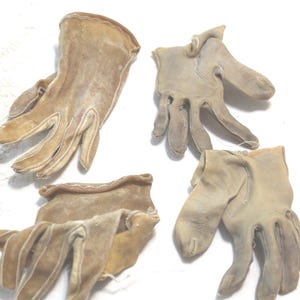 May include: Four small, tan-colored gloves with visible stitching along the seams. The gloves appear to be made of a soft, suede-like material and are arranged in a group. A small, circular metallic coin is positioned at the top center.
