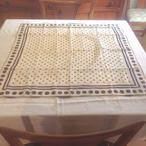 Puede incluir: Un pa&ntilde;uelo cuadrado color crema con un estampado de lunares azul marino. El pa&ntilde;uelo tiene un borde azul marino con una serie de puntos y l&iacute;neas. El pa&ntilde;uelo est&aacute; hecho de una tela ligera y se exhibe sobre un mantel blanco.
