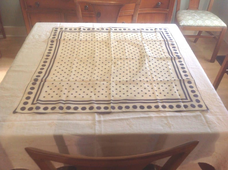 Puede incluir: Un pa&ntilde;uelo cuadrado color crema con un estampado de lunares azul marino. El pa&ntilde;uelo tiene un borde azul marino con una serie de puntos y l&iacute;neas. El pa&ntilde;uelo est&aacute; hecho de una tela ligera y se exhibe sobre un mantel blanco.