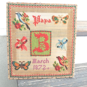 May include: A vintage needlepoint piece with a tan background and a red and green border. The design includes the word "Papa", the letter "B", butterflies, birds, flowers, and the date "March 1872."