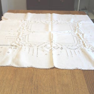 May include: A white, rectangular tablecloth with an intricate cutwork design, featuring geometric patterns and scalloped edges. The cloth is laid out on a wooden table, showcasing its delicate details and texture. This vintage textile is suitable for dining or decorative use.