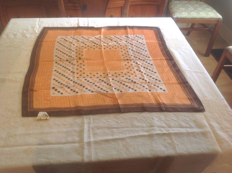 Puede incluir: Un pa&ntilde;uelo cuadrado en tonos naranja y marr&oacute;n. Tiene un borde marr&oacute;n, un centro naranja y un patr&oacute;n de puntos blancos, marrones y negros. El pa&ntilde;uelo est&aacute; hecho de un material ligero y est&aacute; colocado sobre un mantel blanco.