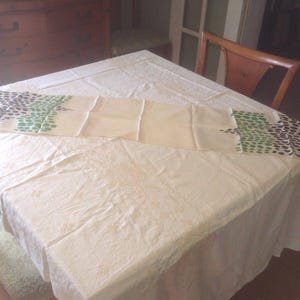 Puede incluir: Un camino de mesa color crema con un patr&oacute;n repetido de puntos verdes y azul oscuro. El camino de mesa est&aacute; colocado sobre un mantel blanco. Una silla y una c&oacute;moda de madera est&aacute;n en el fondo.