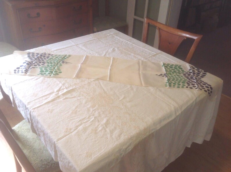 Puede incluir: Un camino de mesa color crema con un patr&oacute;n repetido de puntos verdes y azul oscuro. El camino de mesa est&aacute; colocado sobre un mantel blanco. Una silla y una c&oacute;moda de madera est&aacute;n en el fondo.