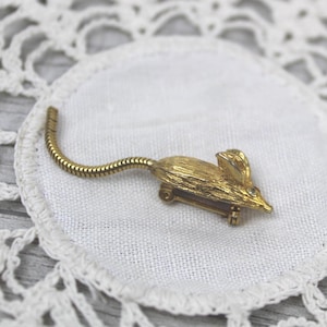 May include: A gold-toned mouse brooch with a textured body and a flexible tail. The mouse has a small, clear gem for an eye. The brooch is set on a white, circular doily.