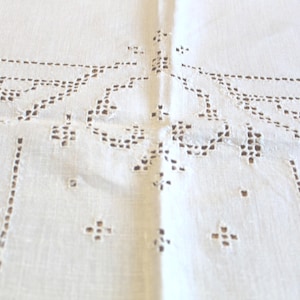 May include: A white textile featuring intricate openwork embroidery with a geometric and scroll pattern. The fabric displays a series of small, square cutouts arranged in linear and curved motifs, creating a delicate, lace-like appearance.