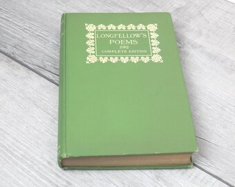 May include: A hardcover book with a green cover. The book's title, "Longfellow's Poems Complete Edition," is displayed in gold lettering within a decorative frame. The book is resting on a light gray wooden surface.