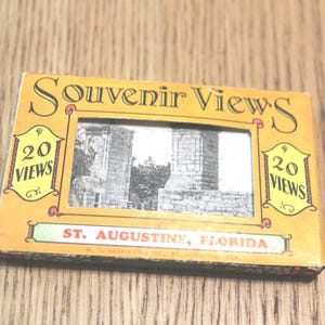 May include: A vintage souvenir box with the title "Souvenir Views" in a decorative font. The box features a black and white image of a building and the text "St. Augustine, Florida". The box is yellow with red and green accents.