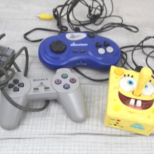 May include: An assortment of retro video game controllers. Includes a blue Intellivision controller, a gray Sony PlayStation controller, and a yellow SpongeBob SquarePants joystick controller, all with attached cables.