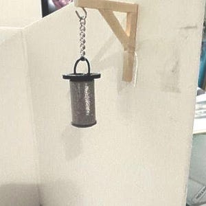 May include: A miniature cylindrical lantern with a metallic mesh body and black top and bottom rims, suspended by a silver-toned metal chain from a light-colored wooden wall-mounted bracket. The lantern features a small handle on the top rim.