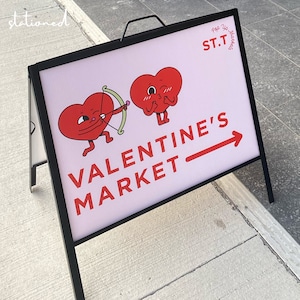 May include: A black metal A-frame sidewalk sign with a white face featuring two red heart characters, one holding a green bow and arrow. The text reads ST.T POP UP MARKETS and VALENTINE'S MARKET with a red arrow pointing to the right.