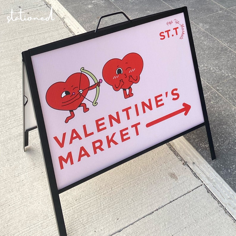 May include: A black metal A-frame sidewalk sign with a white face featuring two red heart characters, one holding a green bow and arrow. The text reads ST.T POP UP MARKETS and VALENTINE'S MARKET with a red arrow pointing to the right.