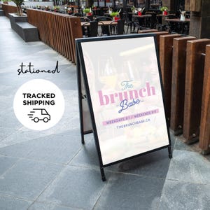 May include: A black metal A-frame sidewalk sign featuring a white poster with the text The brunch Babe, Weekdays 8-1 / Weekends 8-3, and THEBRUNCHBABE.CA. The graphic includes images of two raised wine glasses filled with a golden liquid.