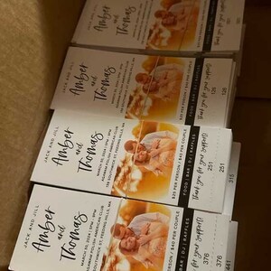Puede incluir: Una pila de tarjetas de invitaci&oacute;n de boda blancas con una foto de una pareja en un campo. El texto en las tarjetas dice "Jack y Jill, Amber y Thomas" e incluye la fecha, la hora y el lugar de la boda. Las tarjetas tambi&eacute;n incluyen informaci&oacute;n sobre el costo de los servicios de comida, bar y DJ.