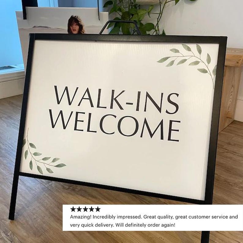 May include: A black metal A-frame sidewalk sign with a white rectangular insert. The sign features the black text WALK-INS WELCOME in a clean sans-serif font, accented by delicate green leaf illustrations in the top right and bottom left corners.