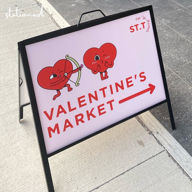 May include: A pink and black sign with a red arrow pointing to the right. The sign says "Valentine's Market" and features two red cartoon hearts with faces. One heart is winking and the other is blowing a kiss. The sign also says "Pop Up Market ST.T."
