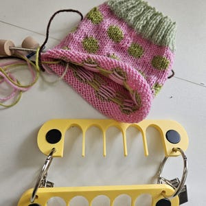 Op de afbeelding: Een gedeeltelijk gebreide muts met roze en groene stippen met bevestigd garen en breinaalden, geplaatst boven twee gele, kamvormige plastic gereedschappen die verbonden zijn door zilveren metalen ringen en zwarte ronde sluitingen.