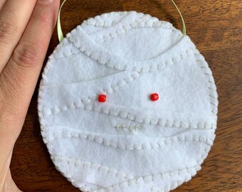 May include: A round, white felt mummy ornament with red bead eyes and gold stitching. The mummy is wrapped in strips of white felt and has a light green ribbon for hanging. A hand is next to the ornament.