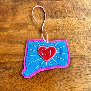 May include: A felt ornament in the shape of Connecticut, with a blue interior and a pink border. A red heart with "CT" is in the center, with gold rays emanating from it. A pink ribbon is attached for hanging.