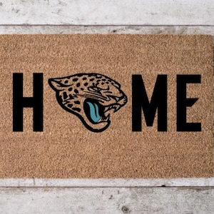 May include: A rectangular doormat with the word "HOME" in bold black letters. The "O" is replaced by a detailed illustration of a jaguar's head, featuring black spots, a blue tongue, and a fierce expression. The mat is a natural tan color.