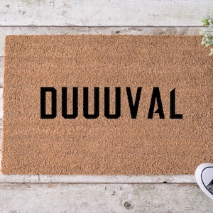May include: A rectangular brown coir doormat featuring the word DUUUVAL printed in bold black capital letters. The mat has a coarse, fibrous texture and is positioned on a light-colored wooden surface with a small green plant and sneakers.