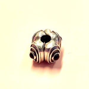 Pu&ograve; includere: Una perlina argentata dal design unico. La perlina presenta una serie di linee curve e concentriche e piccoli accenti scuri. Ha un'apertura centrale e viene probabilmente utilizzata per la creazione di gioielli o come elemento decorativo.