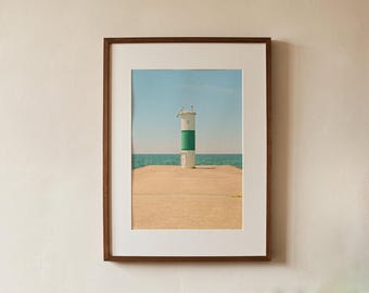 May include: A framed photograph featuring a white and green cylindrical lighthouse standing on a concrete pier extending into the ocean under a clear blue sky. The print is encased in a dark brown wooden frame with a wide white mat border.