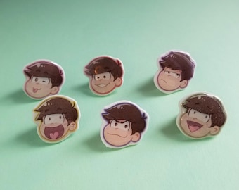 May include: Six colorful cartoon character pins with different facial expressions. The pins are round and feature characters with brown hair and white eyes.