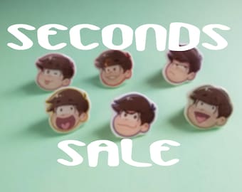 May include: A collection of six cartoon character pins featuring different expressions. The pins are on a light green background with the text "SECONDS SALE" in white.