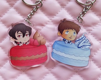 May include: Two keychains featuring cartoon characters on top of cake-like desserts. The keychain on the left features a red cake with chocolate and a strawberry on top. The keychain on the right features a blue cake with white frosting and a swirl of blue frosting on top.