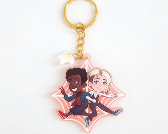 May include: A pink and white web-shaped keychain with a gold chain and a star charm. Two cartoon figures of Spider-Man characters are on the keychain, one is black and the other is white.