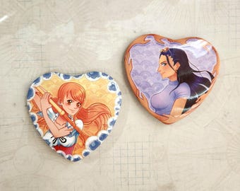 May include: Two heart-shaped buttons with anime characters. The first button features a woman with orange hair holding a sword, wearing a blue and white dress. The second button features a woman with long black hair wearing a purple shirt.