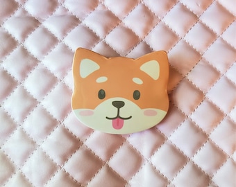 May include: A cartoon Shiba Inu dog face with a pink tongue sticking out. The dog is orange and white with black eyes. The background is a pink quilted fabric.
