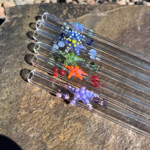 May include: A collection of six clear glass straws, each adorned with colorful, handcrafted starfish designs. The starfish are in various colors, including blue, yellow, orange, green, and purple, with small glass beads. The straws are arranged diagonally on a stone surface.