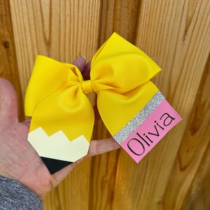 May include: A yellow pencil-shaped hair bow with a pink and silver glitter tag that says "Olivia".