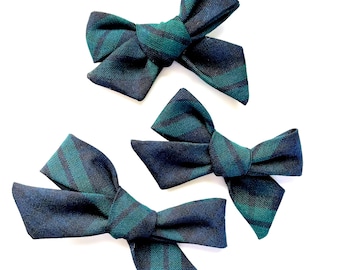 May include: Three plaid hair bows in shades of green and navy blue. The bows are tied in a classic bow shape and are ready to add a touch of style to any outfit.