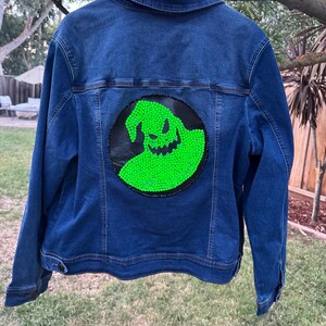 May include: A dark blue denim jacket with a large, circular patch on the back. The patch features a neon green, stylized character design on a black background. The jacket is hanging on a white hanger.