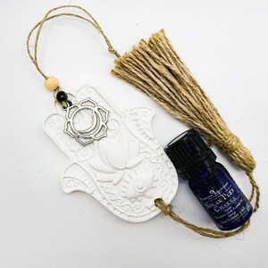 May include: White ceramic hamsa hand charm with a silver lotus flower and a silver circle with a crescent moon design. The charm is hanging from a brown twine string with a brown tassel. A small blue bottle of essential oil with a black cap and a white label that reads "Solar Plexus Chakra" is also included.